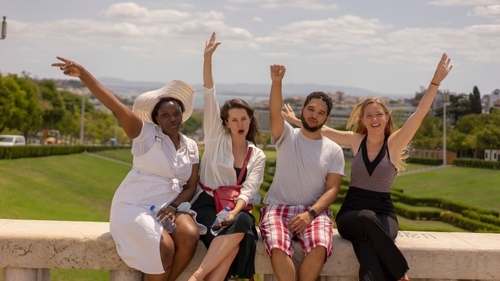 Tracy, Kate, Allan and Bibi sitting on a wall in front of a scenic view of a park. They're cheering with their hands in the air.