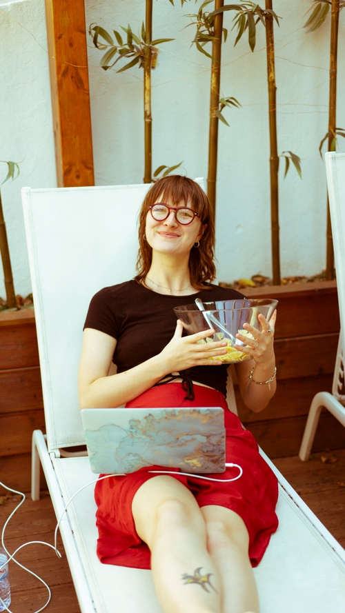 A woman on a reclining chair holding a bowl of pasta, with a laptop on her lap.