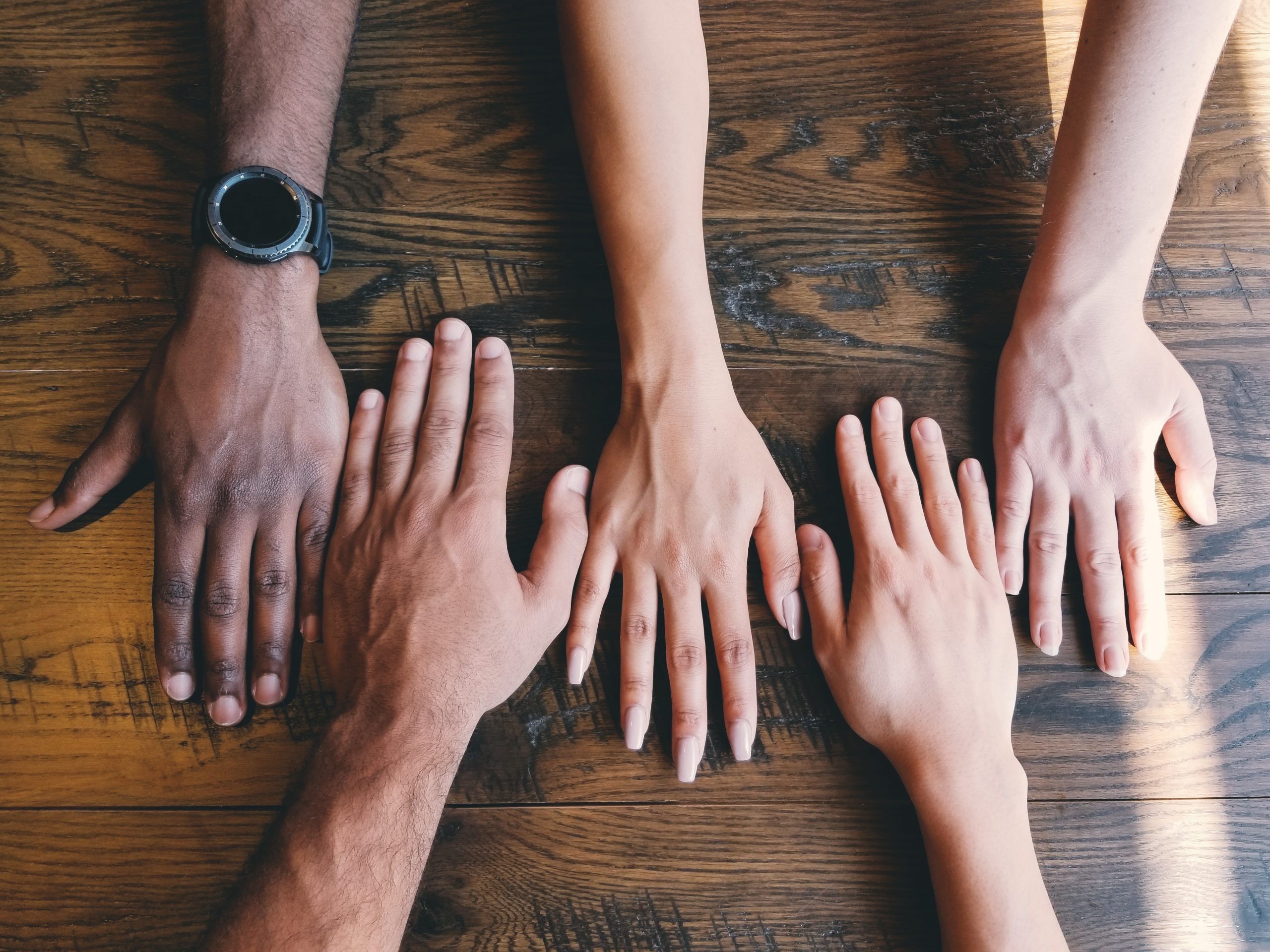 5 hands palm down on a wooden table, all of different skin tones.