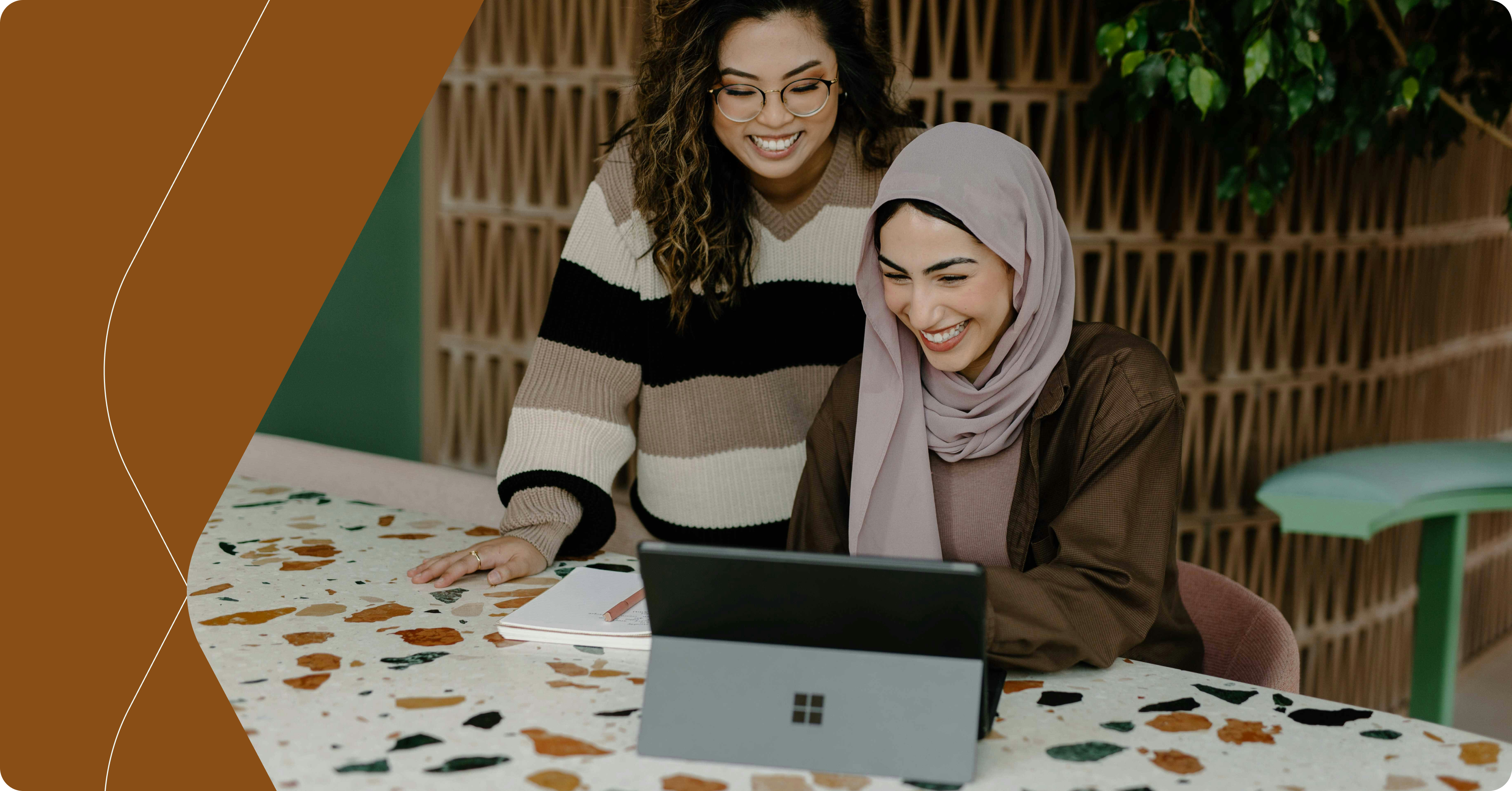 “Two businesswomen collaborating on a laptop: A smiling woman in a hijab and another woman with glasses work together at a modern, terrazzo-style table, highlighting inclusivity and teamwork in a professional setting.”